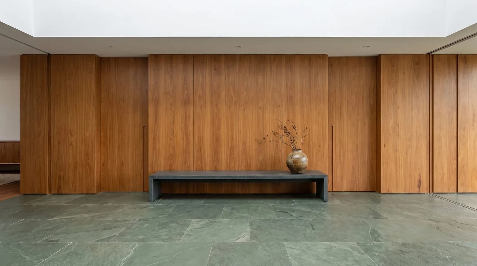 Banco minimalista escuro sobre piso de pedra verde diante de parede de madeira, com vaso decorativo e galhos secos.